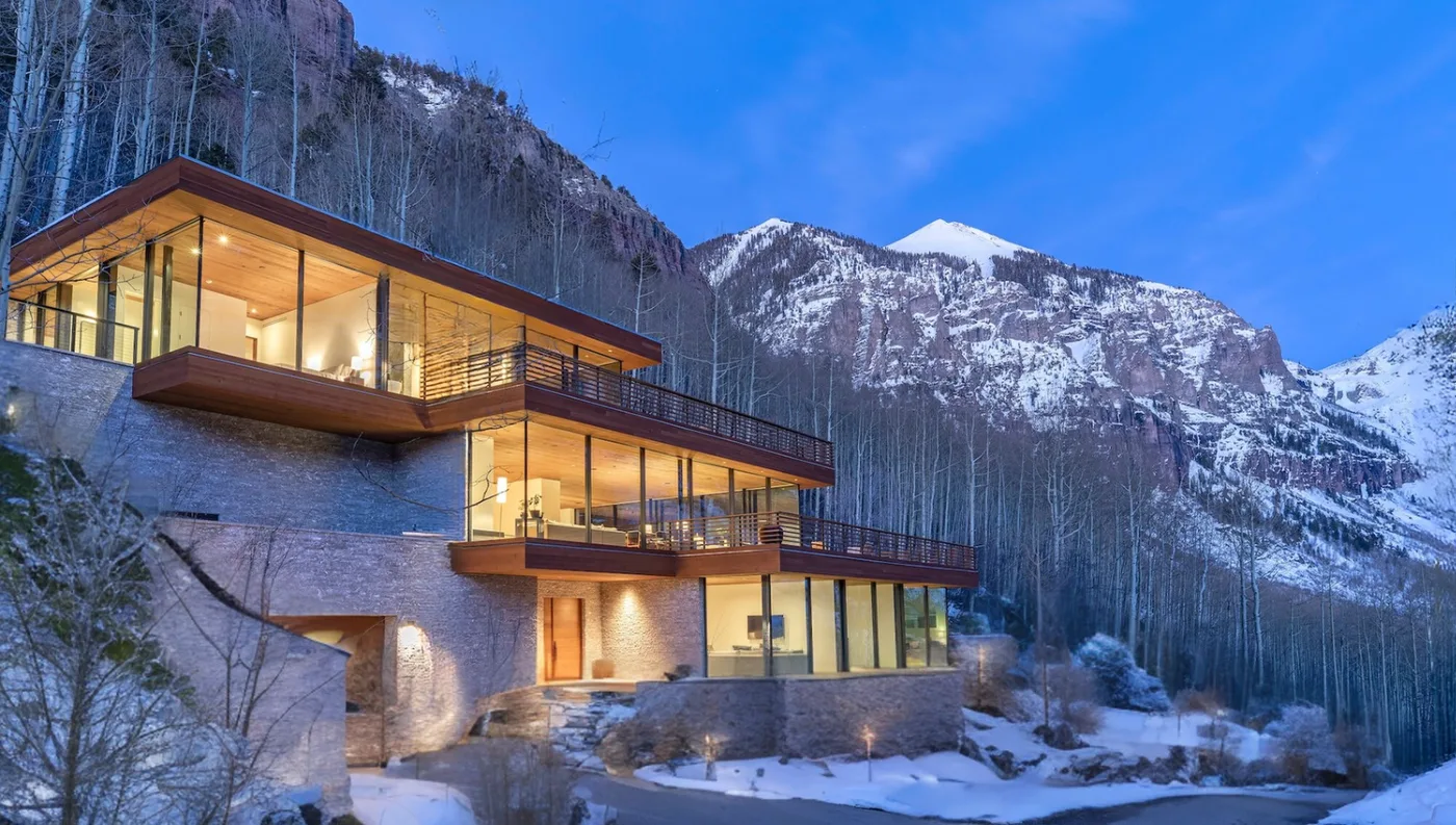 Una casa moderna en la ladera, con ventanas iluminadas, se encuentra bajo montañas nevadas.