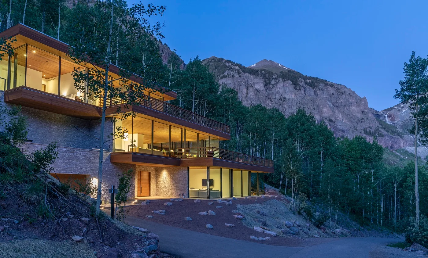 Un edificio moderno con ventanas iluminadas se encuentra entre árboles bajo una montaña.