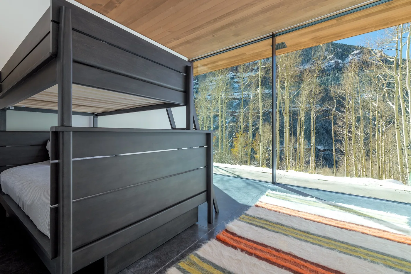Un dormitorio con una litera oscura junto a ventanales con vistas a árboles.