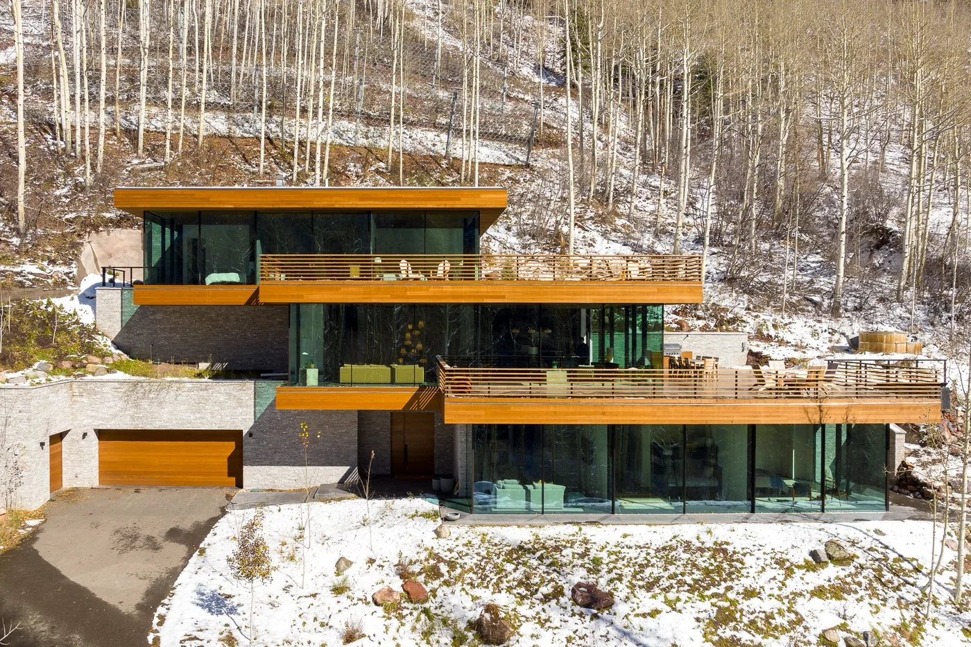 Una casa moderna con fachada de vidrio se encuentra en una ladera boscosa y nevada.