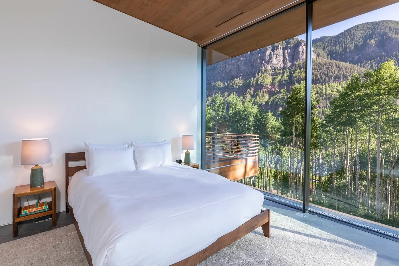 Un dormitorio con una cama blanca junto a ventanales con vistas a una montaña boscosa.