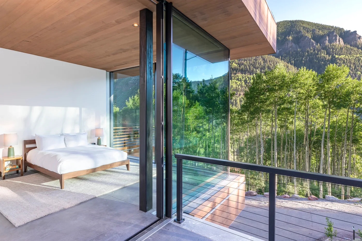 Un dormitorio da a un balcón acristalado con vistas a un paisaje montañoso y arbolado.