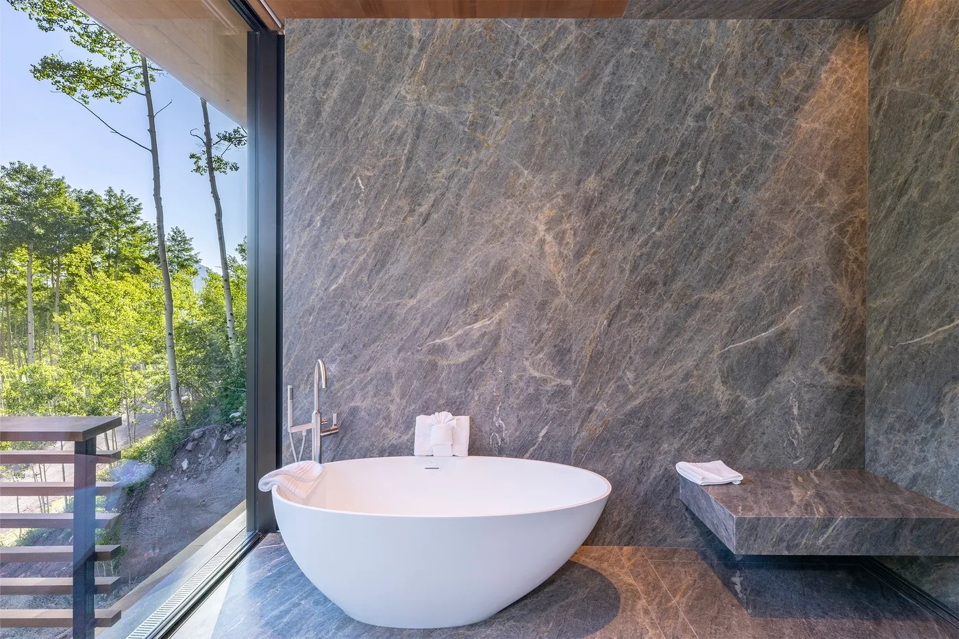 Una bañera exenta está junto a una ventana de piso a techo con vista a los árboles.