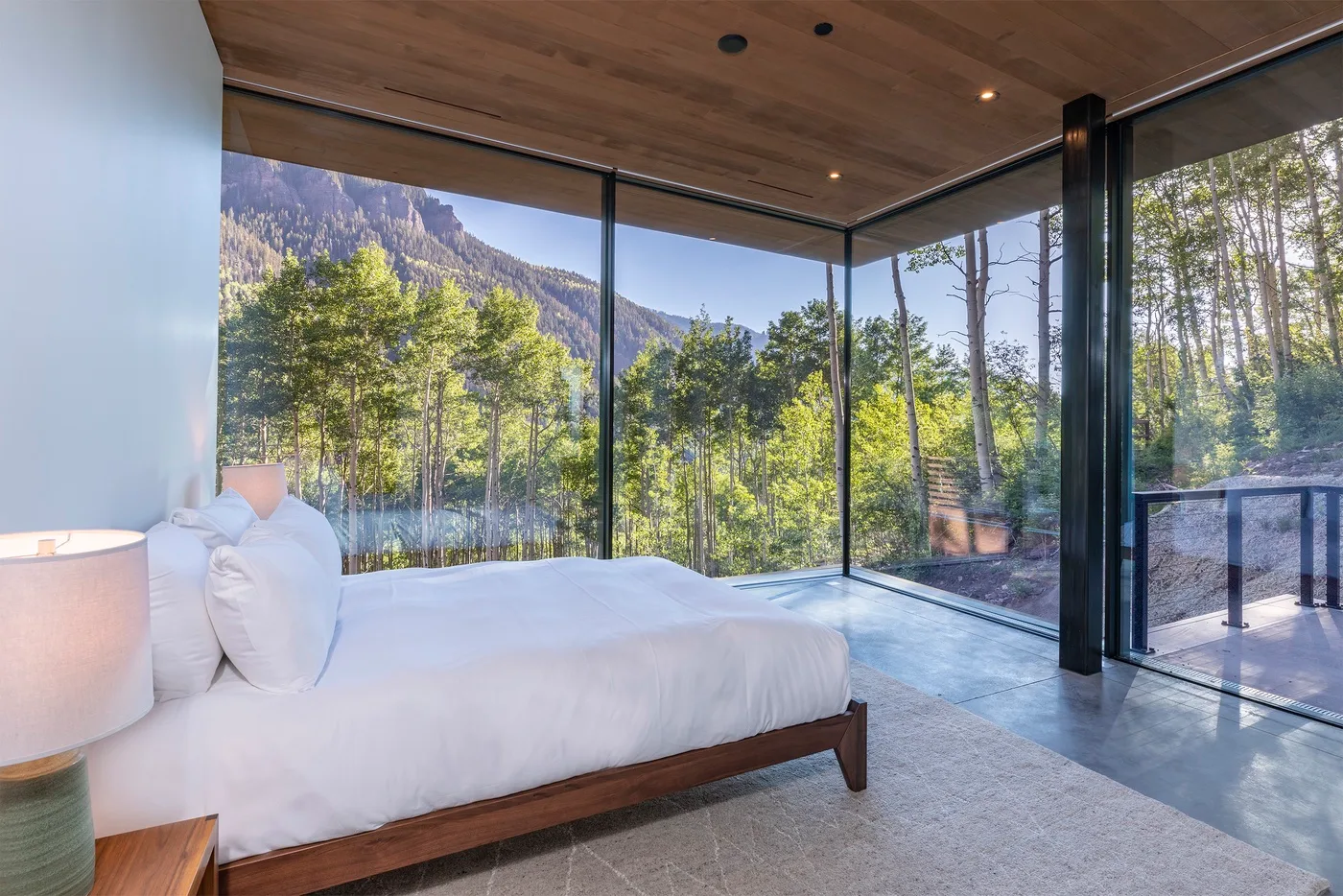 Un dormitorio con una cama baja junto a ventanales con vistas a árboles y montañas.
