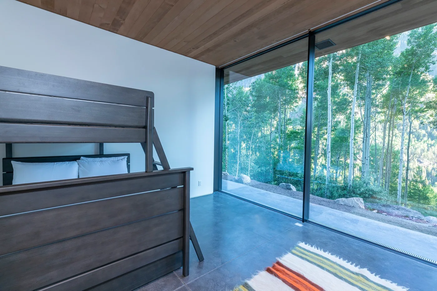 Dormitorio con una litera de madera oscura junto a ventanales con vistas a los árboles.