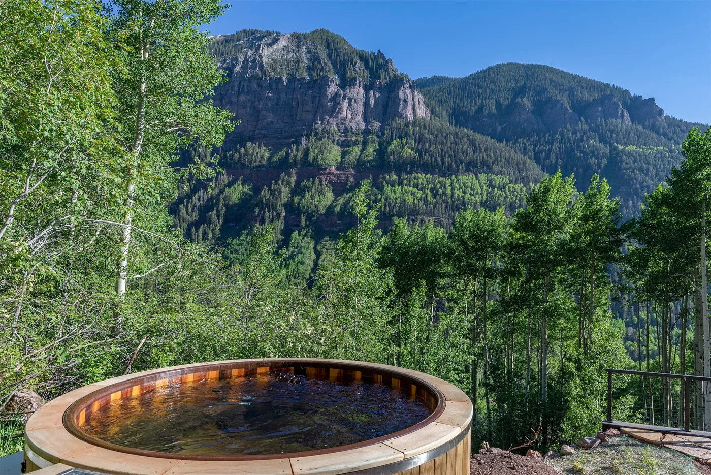 Una bañera de hidromasaje redonda al aire libre está entre árboles, con una montaña al fondo.