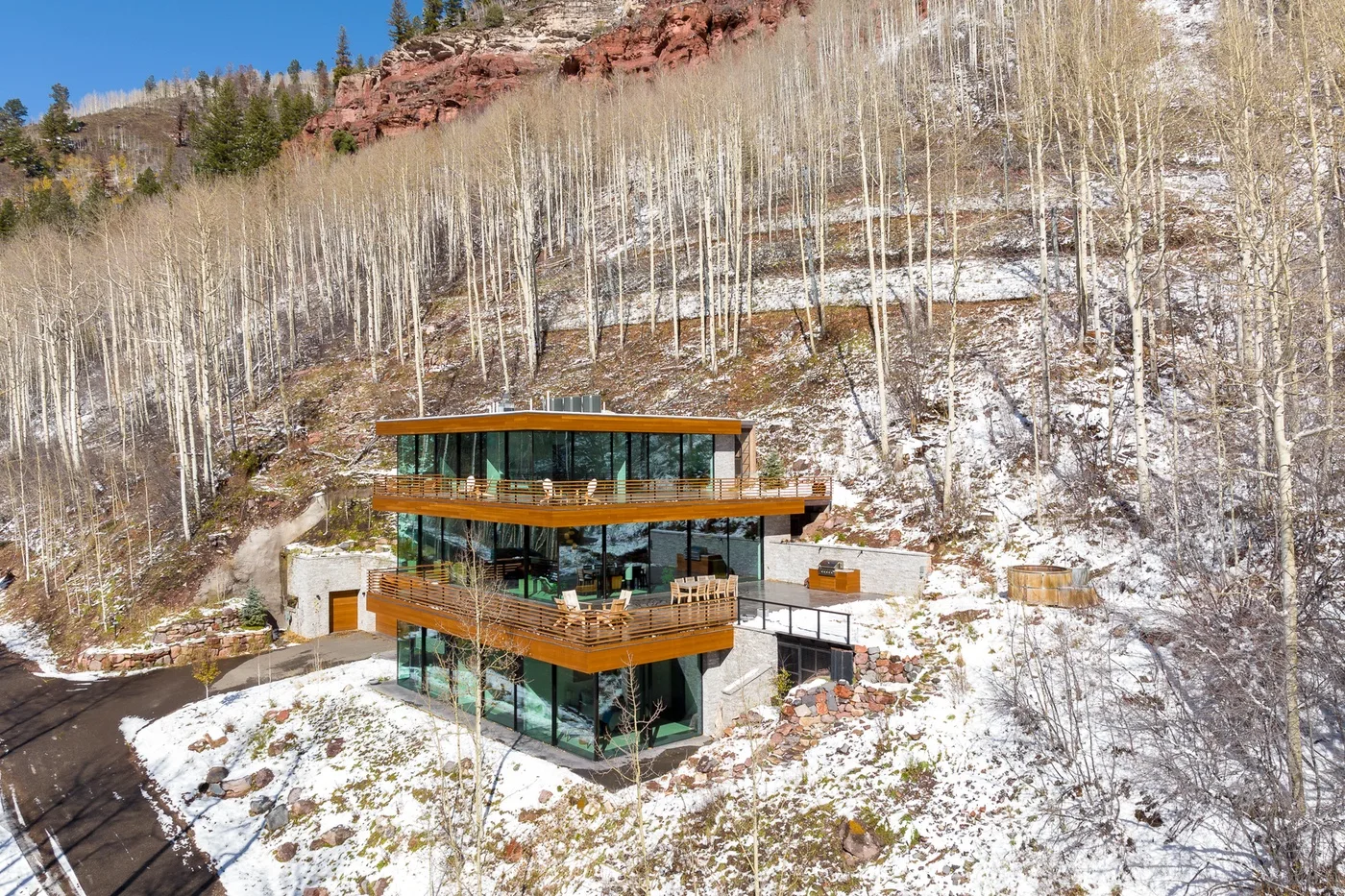 Una casa moderna de cristal se encuentra en una ladera nevada entre árboles sin hojas.