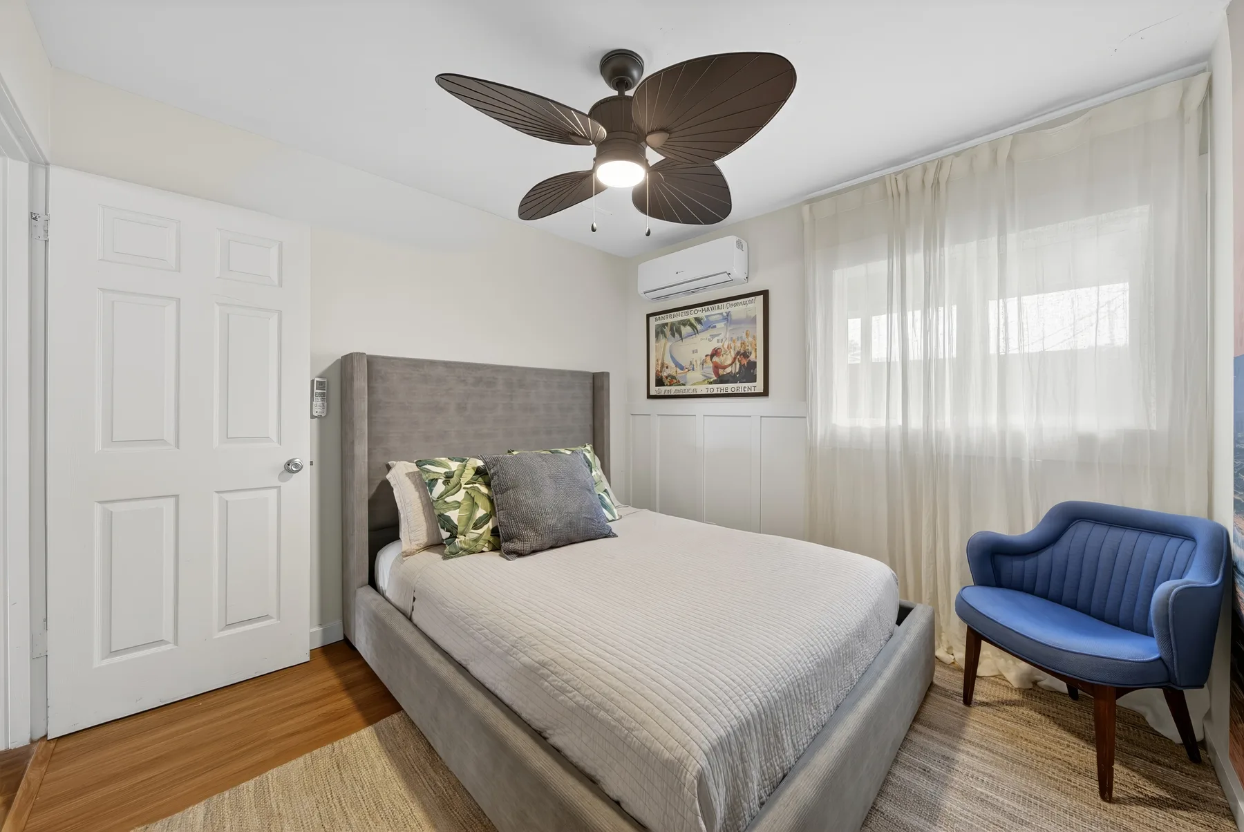 Un dormitorio con una cama bien tendida, un sillón azul y cortinas translúcidas.