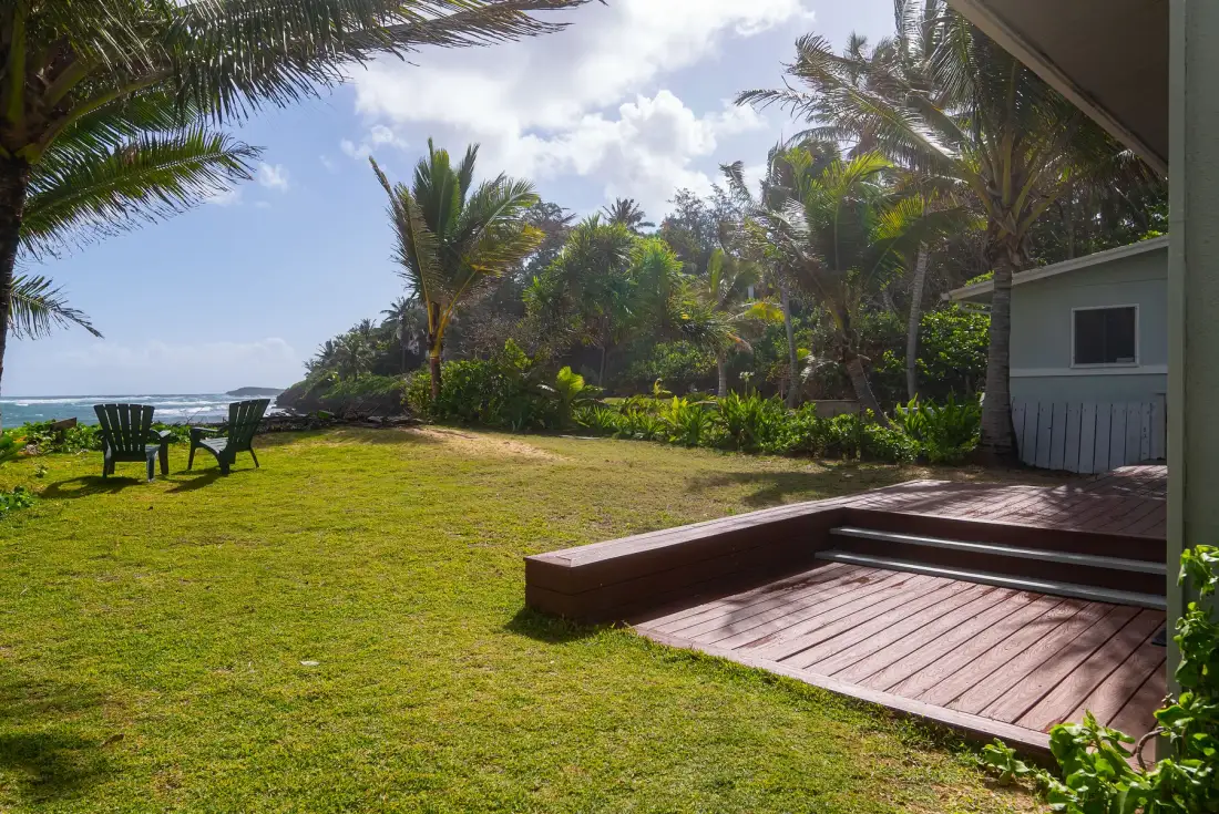 Zona exterior con césped, una terraza de madera, plantas tropicales y vistas al mar.