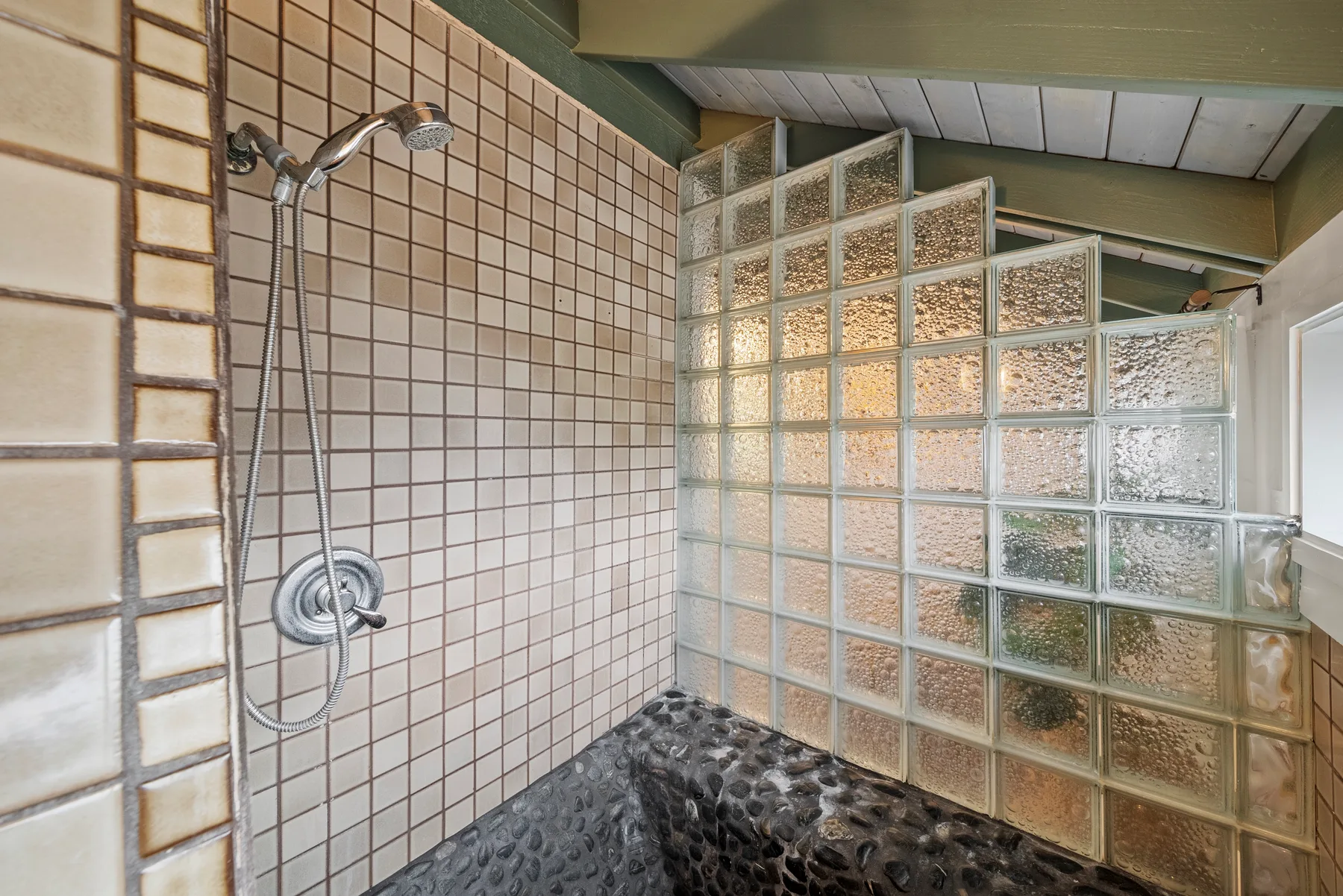 Una zona de ducha alicatada con ducha de mano y una pared de bloques de vidrio.