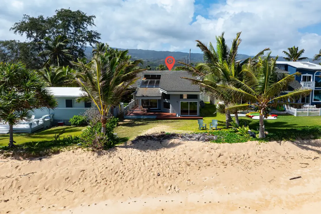 Una casa junto a la playa está detrás de una orilla arenosa, con palmeras y colinas al fondo.