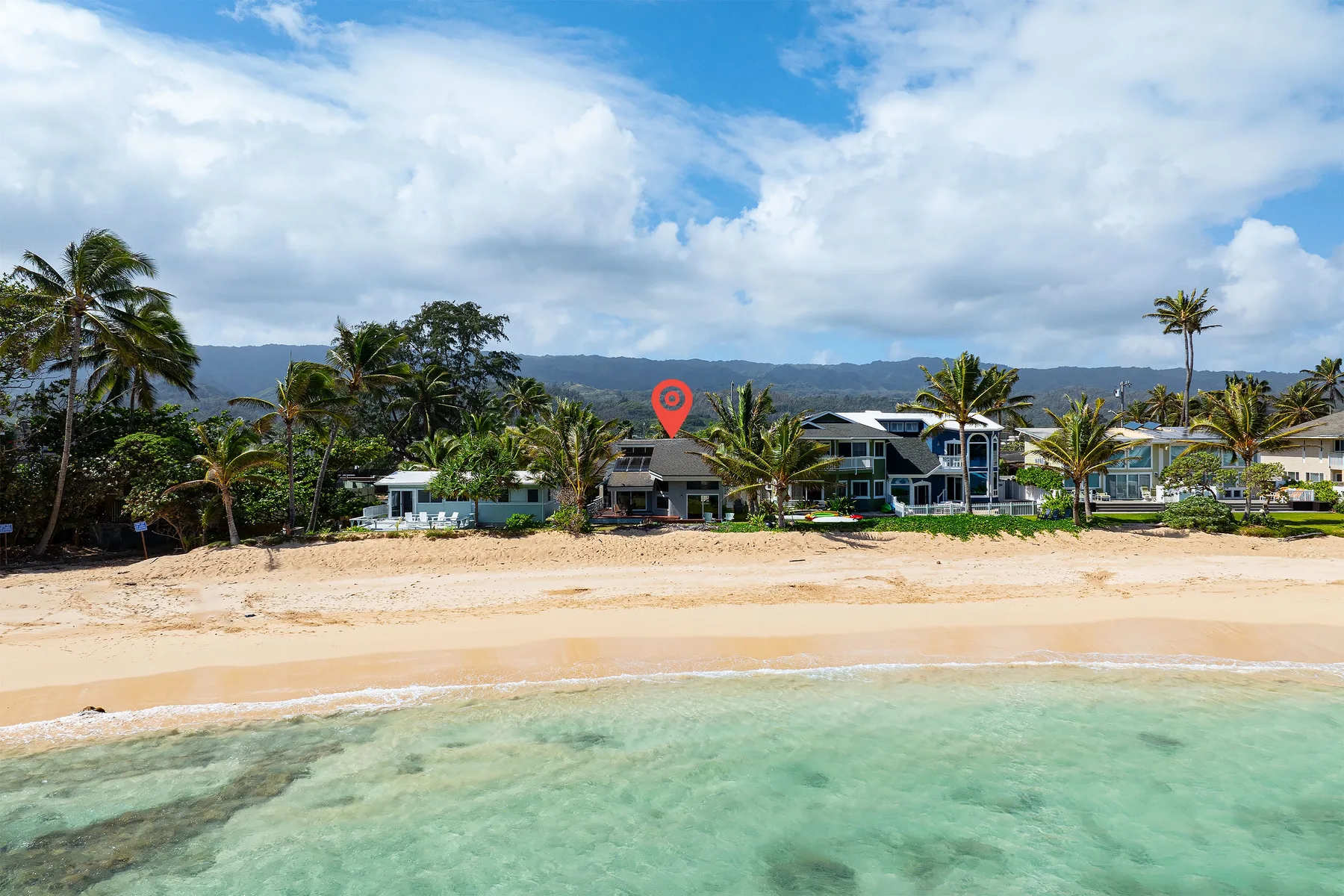 Una playa de arena y aguas claras y poco profundas bordean varios edificios y palmeras.