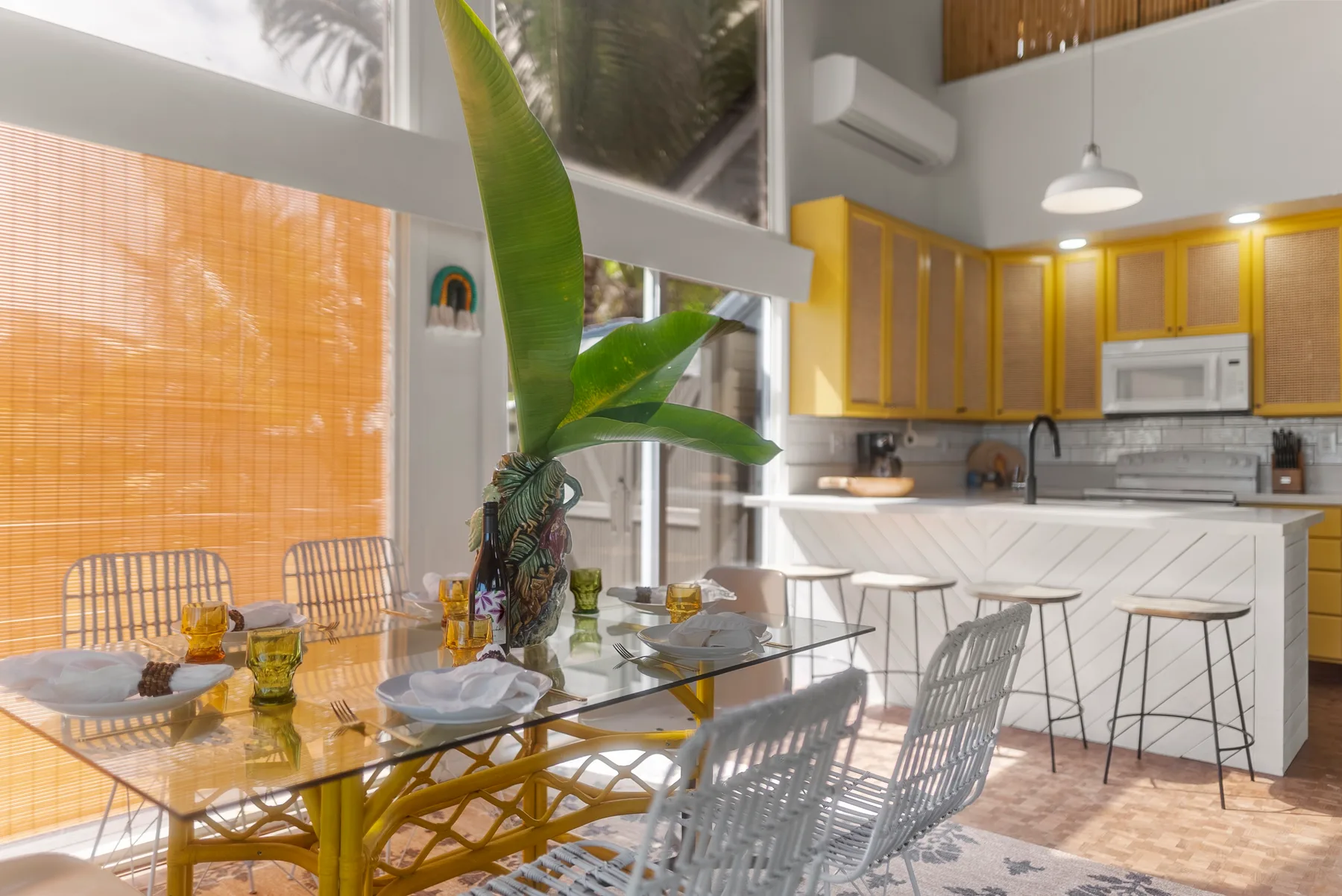Un comedor luminoso junto a una cocina abierta, con mesas de vidrio y asientos tejidos.
