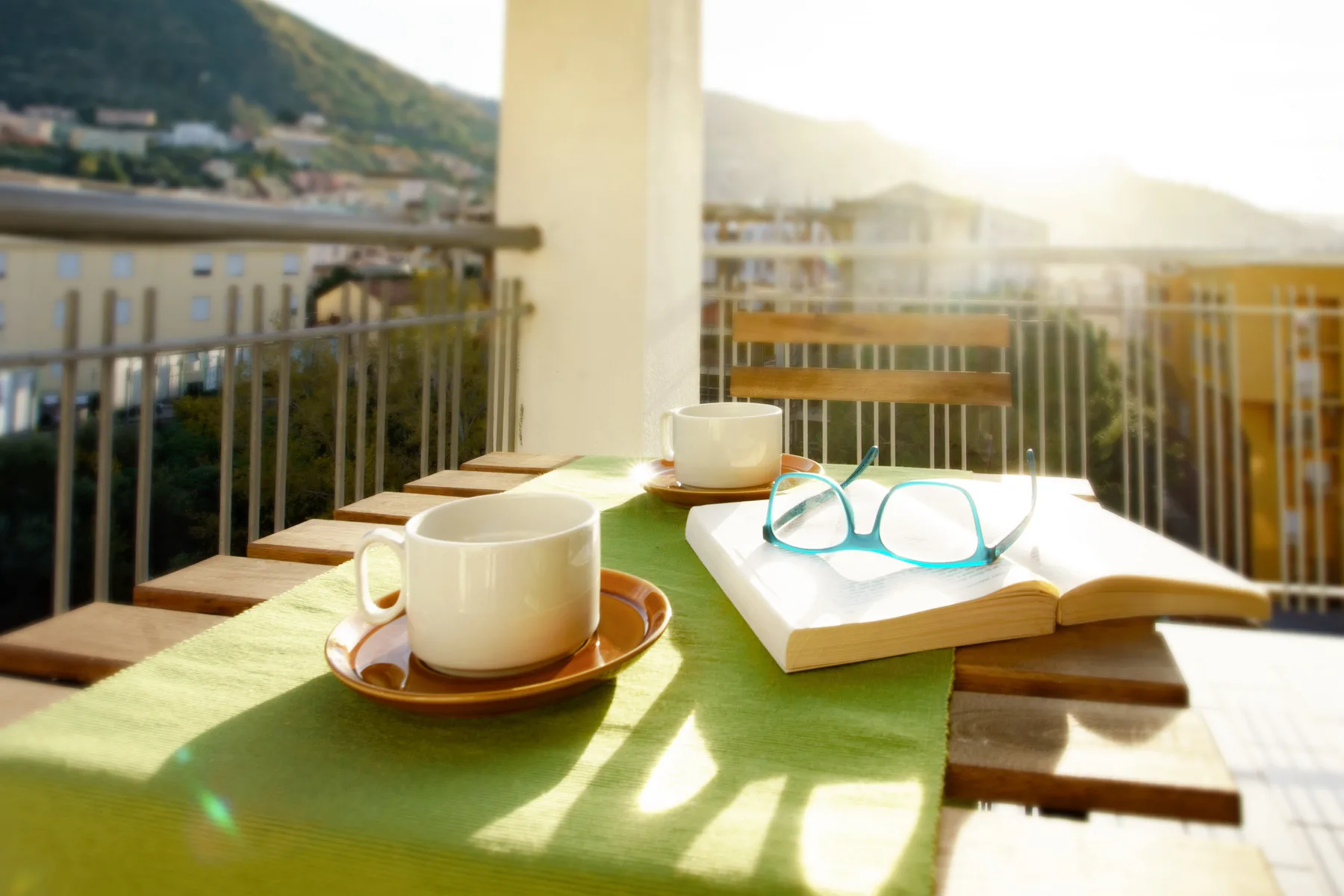 A sunlit balcony table holds cups, eyeglasses, and an open book.