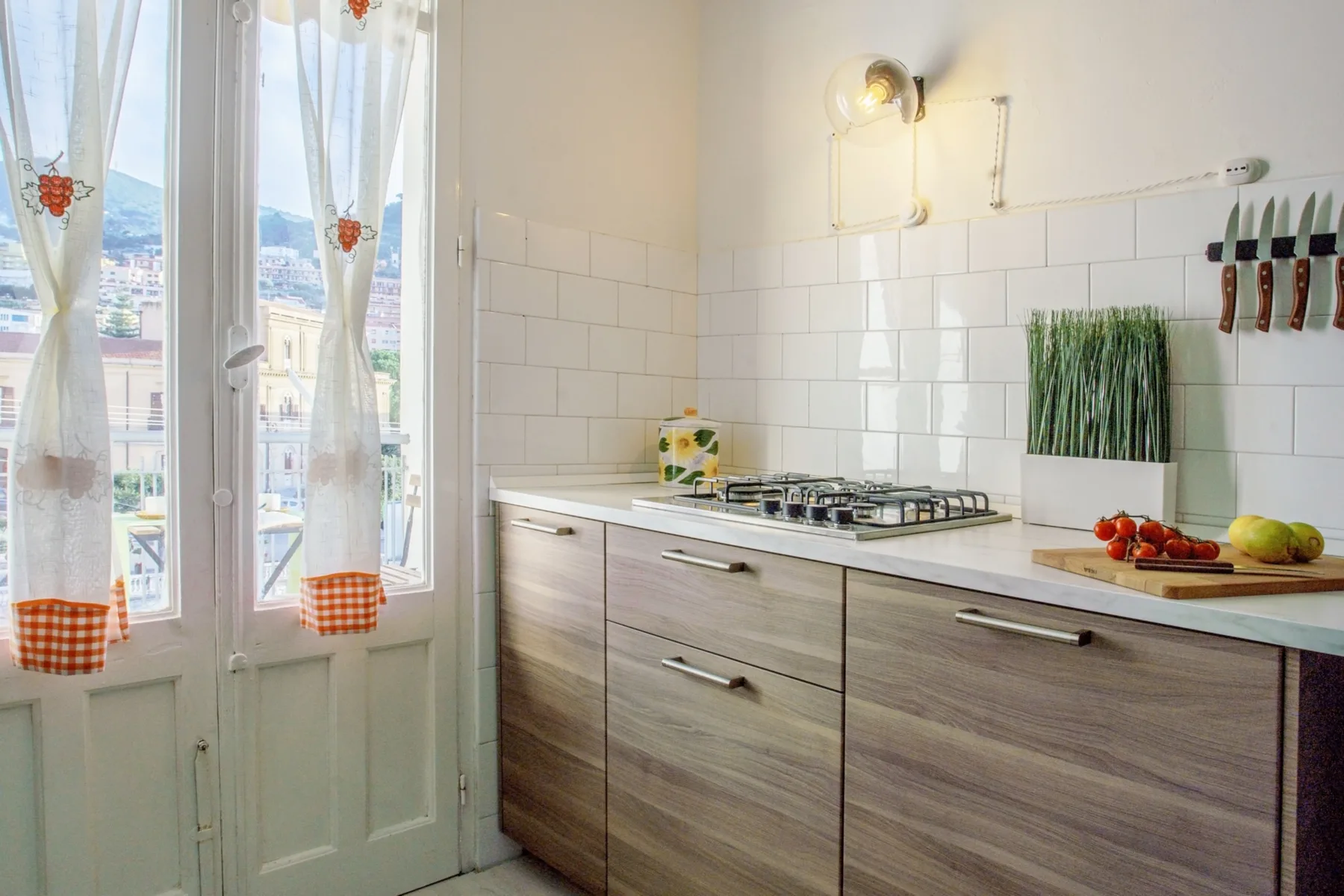 A bright kitchen with a cooktop, wood-tone cabinets, and a window overlooking the city.