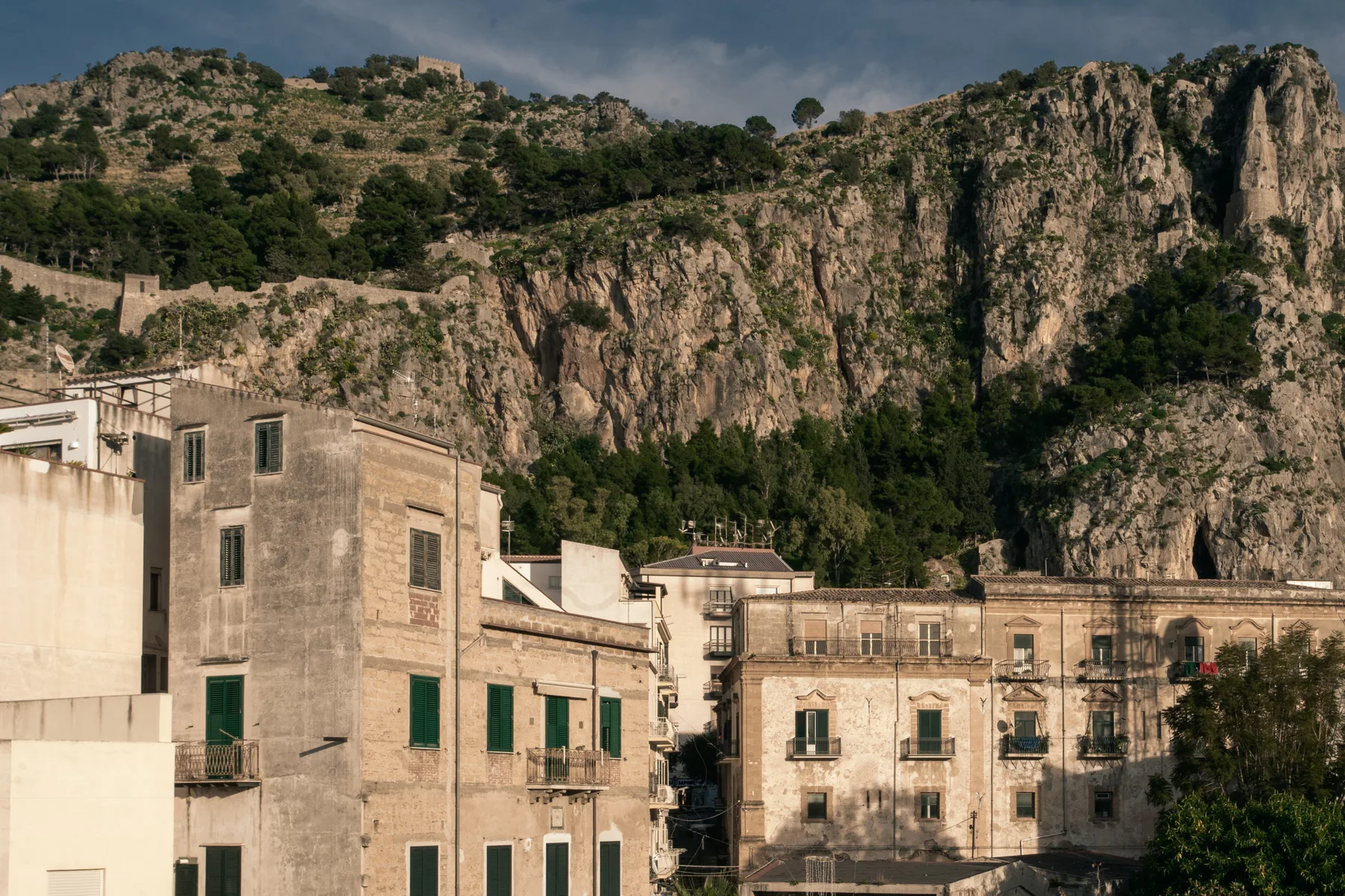 A hillside town with stone buildings beneath a rugged, tree-covered mountain.