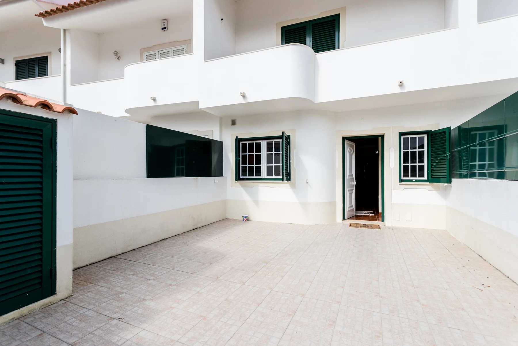 A tiled outdoor courtyard sits beside a white building with green shutters.