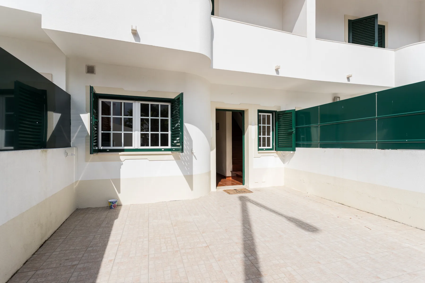 A sunlit courtyard with paved ground, white walls, and green shutters.