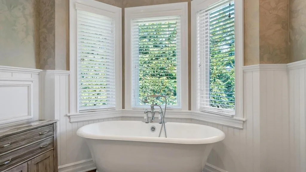 A bathroom with a freestanding bathtub beneath three shuttered windows.
