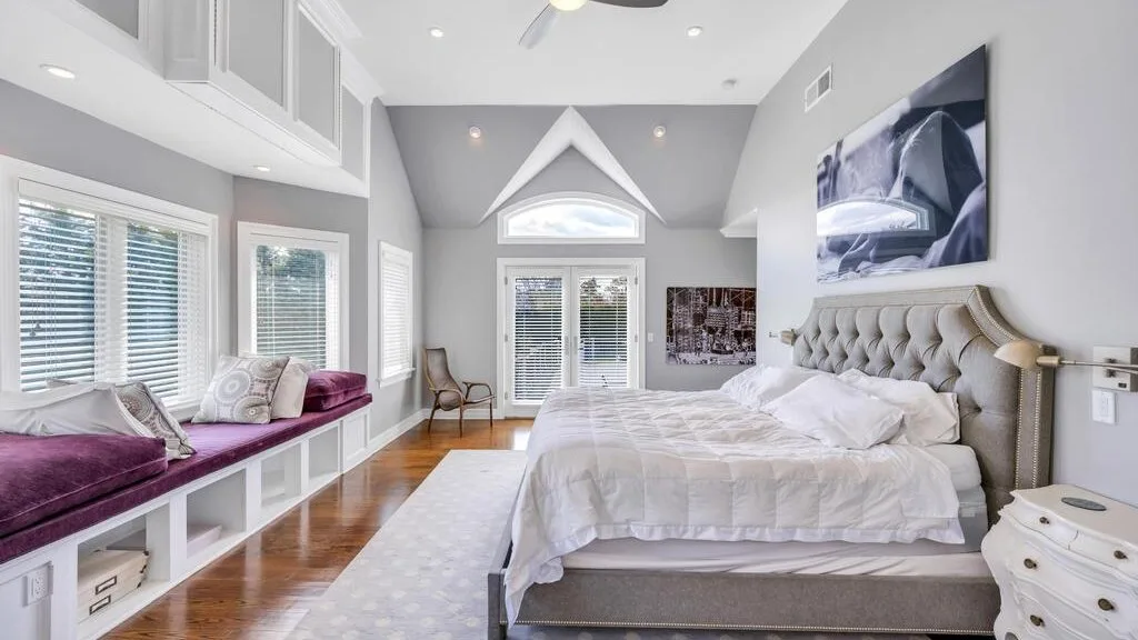 A bright bedroom with a large bed and a window seat beneath wide windows.