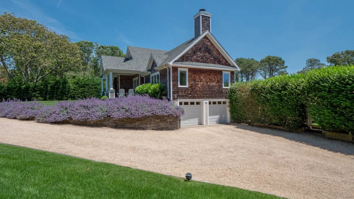 A shingled house stands beside a broad gravel driveway and landscaped garden.