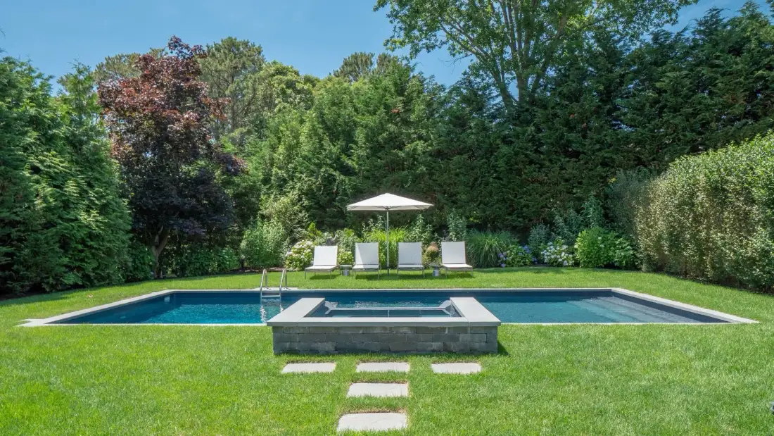 A garden swimming pool sits on a lawn, surrounded by trees and shrubs.