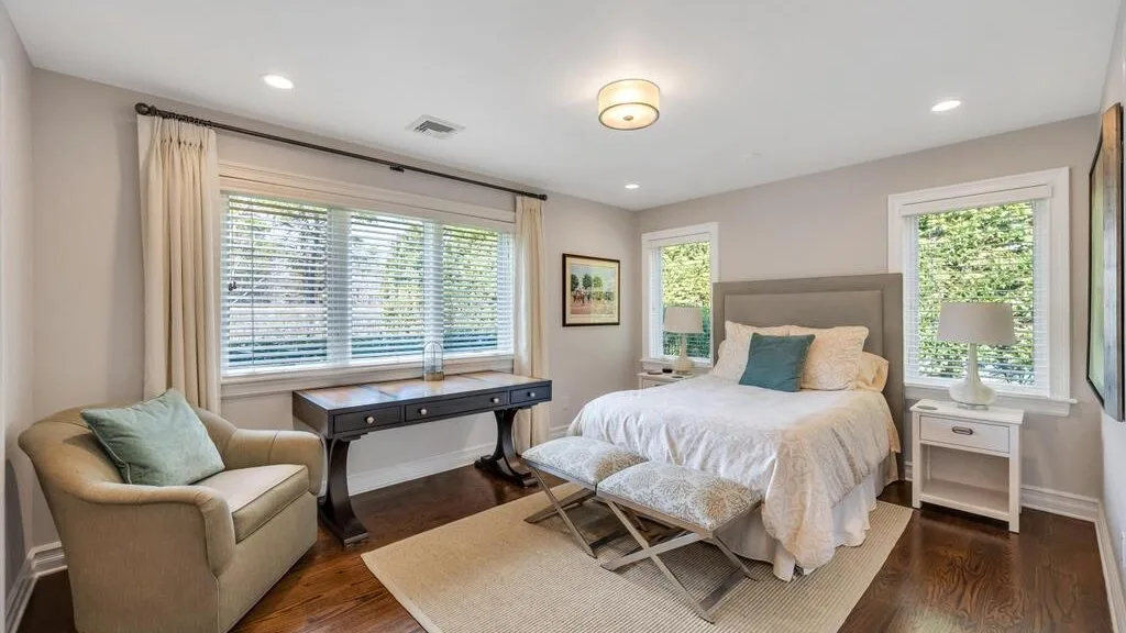 A bedroom with a bed, an upholstered armchair, and a desk beneath the window.