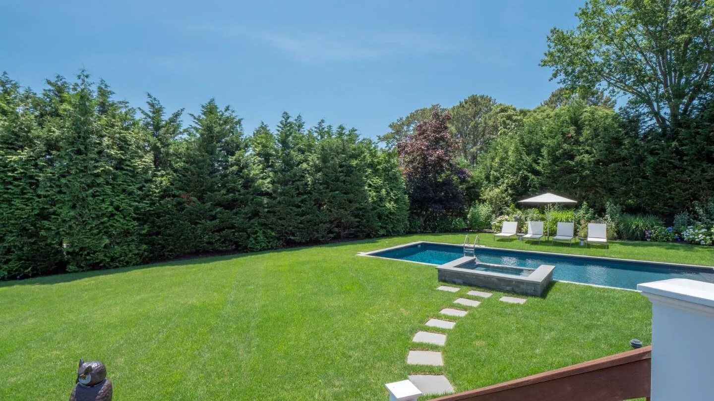 A backyard lawn surrounds a swimming pool, with stepping stones and a row of trees.