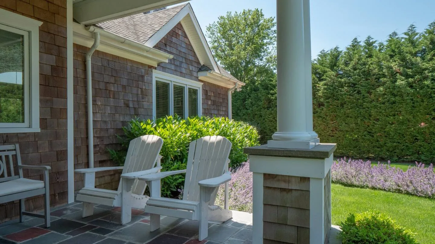 A covered porch with two outdoor seats overlooks a green lawn and flowering plants.