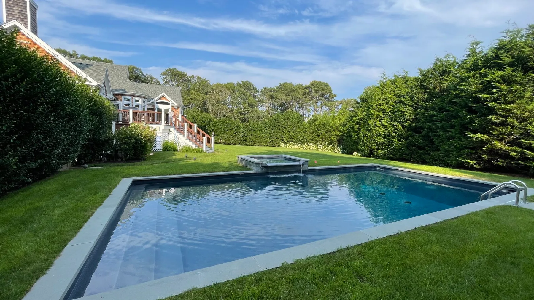 A backyard swimming pool sits beside a lawn, with a house in the background.