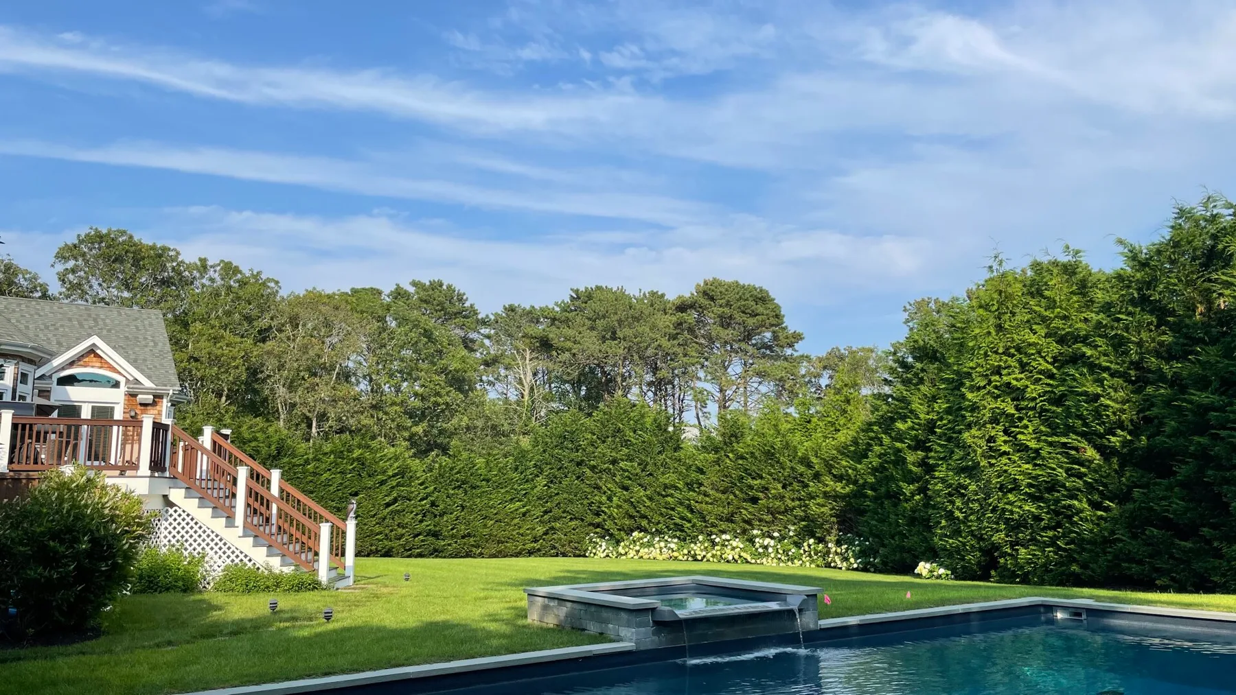 A swimming pool borders a green lawn beside a house and tall hedges.