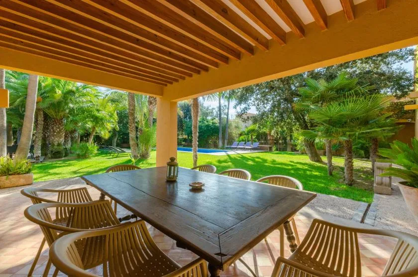 Un comedor exterior cubierto da a un jardín verde con árboles y césped.