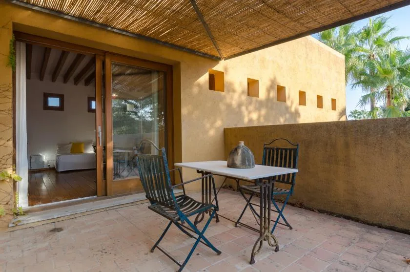Patio sombreado con una mesa y sillas junto a una puerta corredera de vidrio abierta.