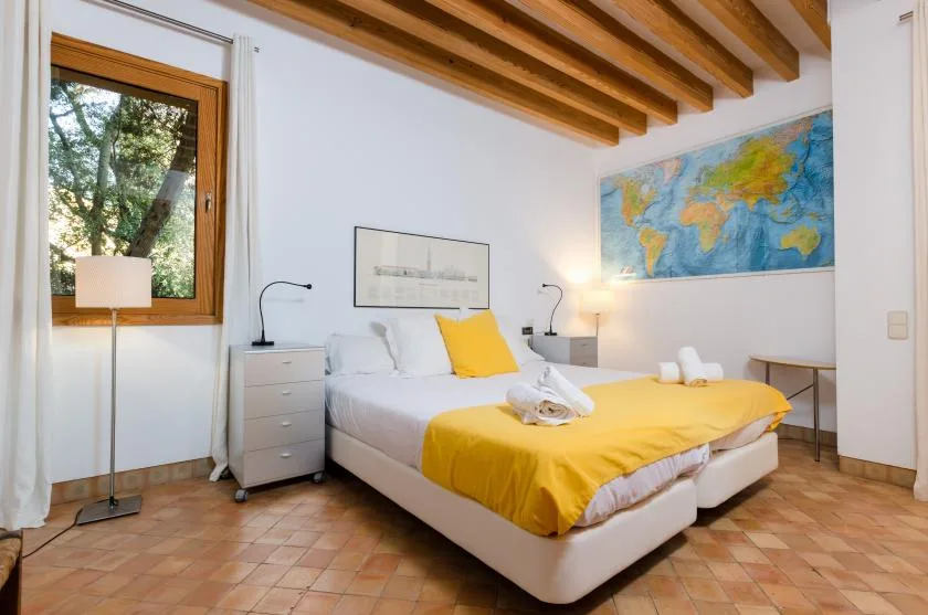 Dormitorio con una cama vestida de blanco y amarillo bajo vigas de madera a la vista.