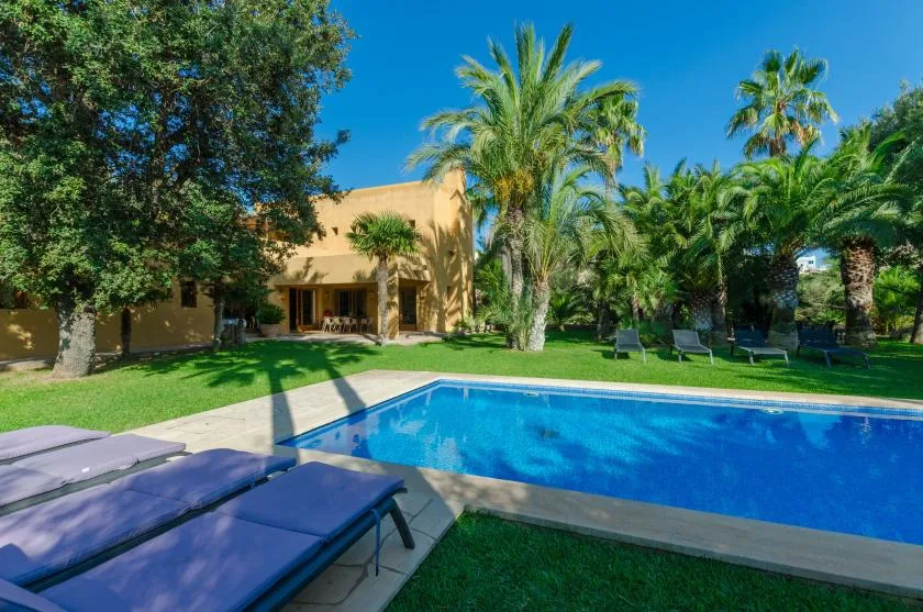 Una piscina está en un jardín soleado junto a un edificio color arena y palmeras.