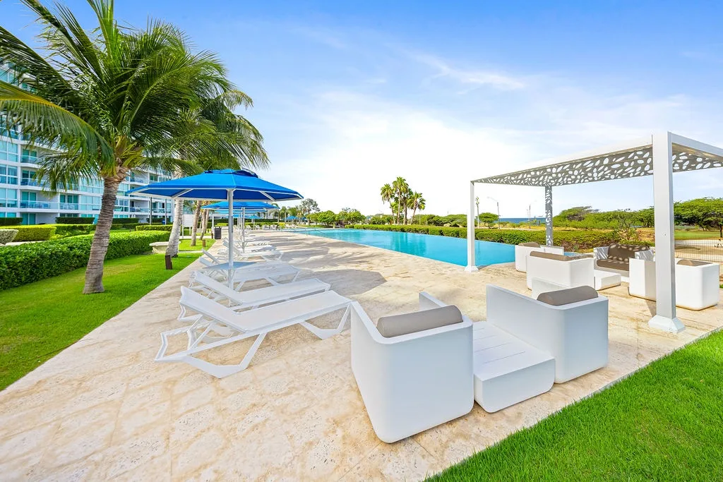 Luxurious pool deck with modern loungers, blue umbrellas, and elegant pergola overlooking tropical landscaping and resort buildings.