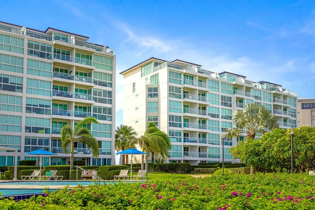 Modern apartment complex with balconies surrounded by tropical gardens, pool, and lush landscaping under bright blue skies.