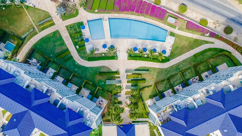 Aerial view shows the property's blue-roofed buildings surrounding a central curved pool with loungers, lush tropical gardens, palm trees, and vibrant flowering plants throughout the landscaped grounds.