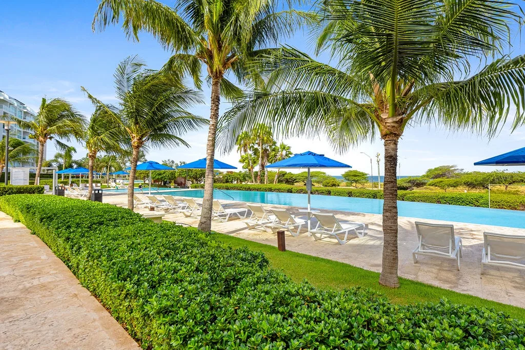 Resort pool area with palm trees, lounge chairs, and blue umbrellas under clear tropical skies.