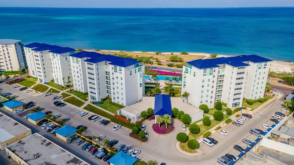 Beachfront property complex situated directly along turquoise waters, surrounded by tropical landscaping and convenient parking areas, with sandy shores and coastal scenery nearby.