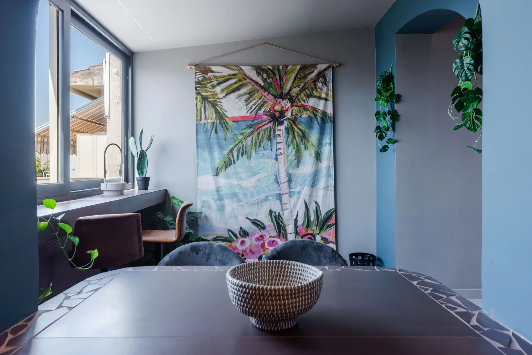 A dining area with a table, woven bowl, and tropical palm artwork.