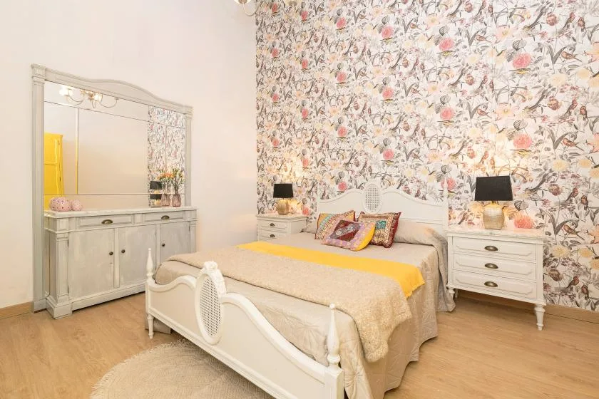 A bedroom with a white bed and floral wallpaper behind it.