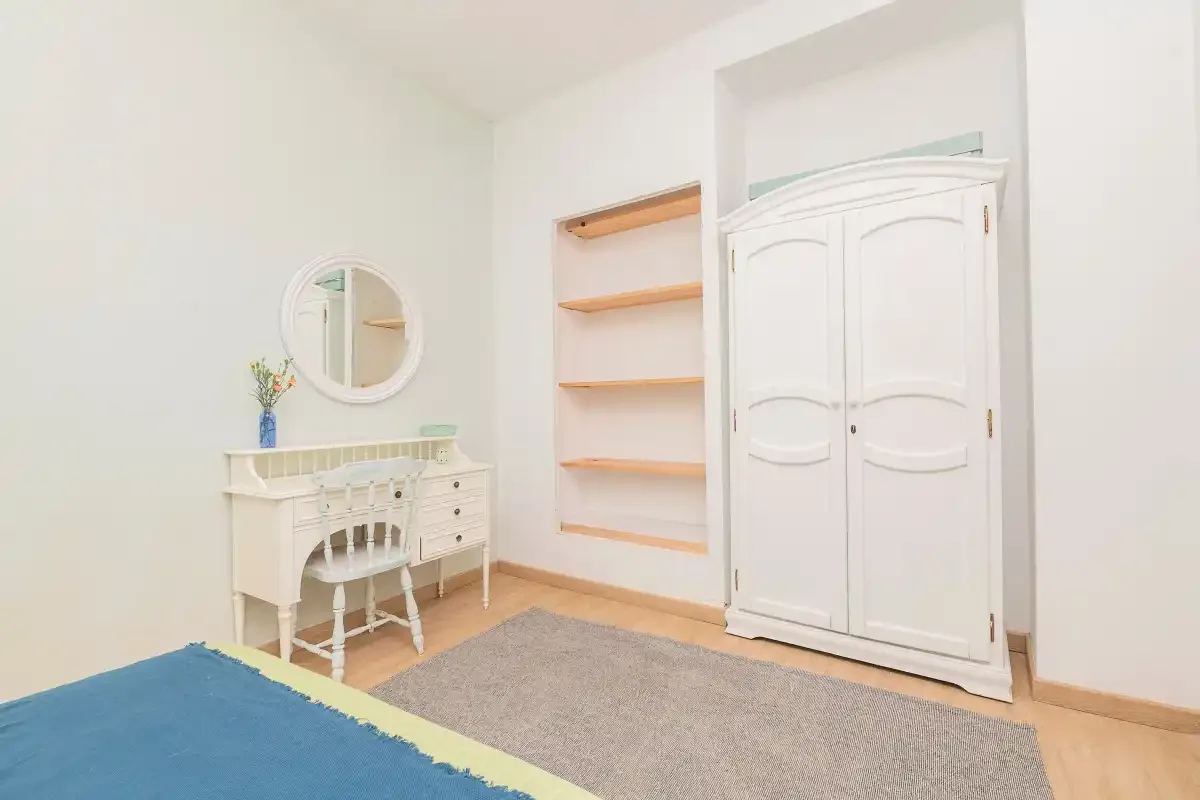 A bright bedroom with a white wardrobe, vanity, and open shelving.