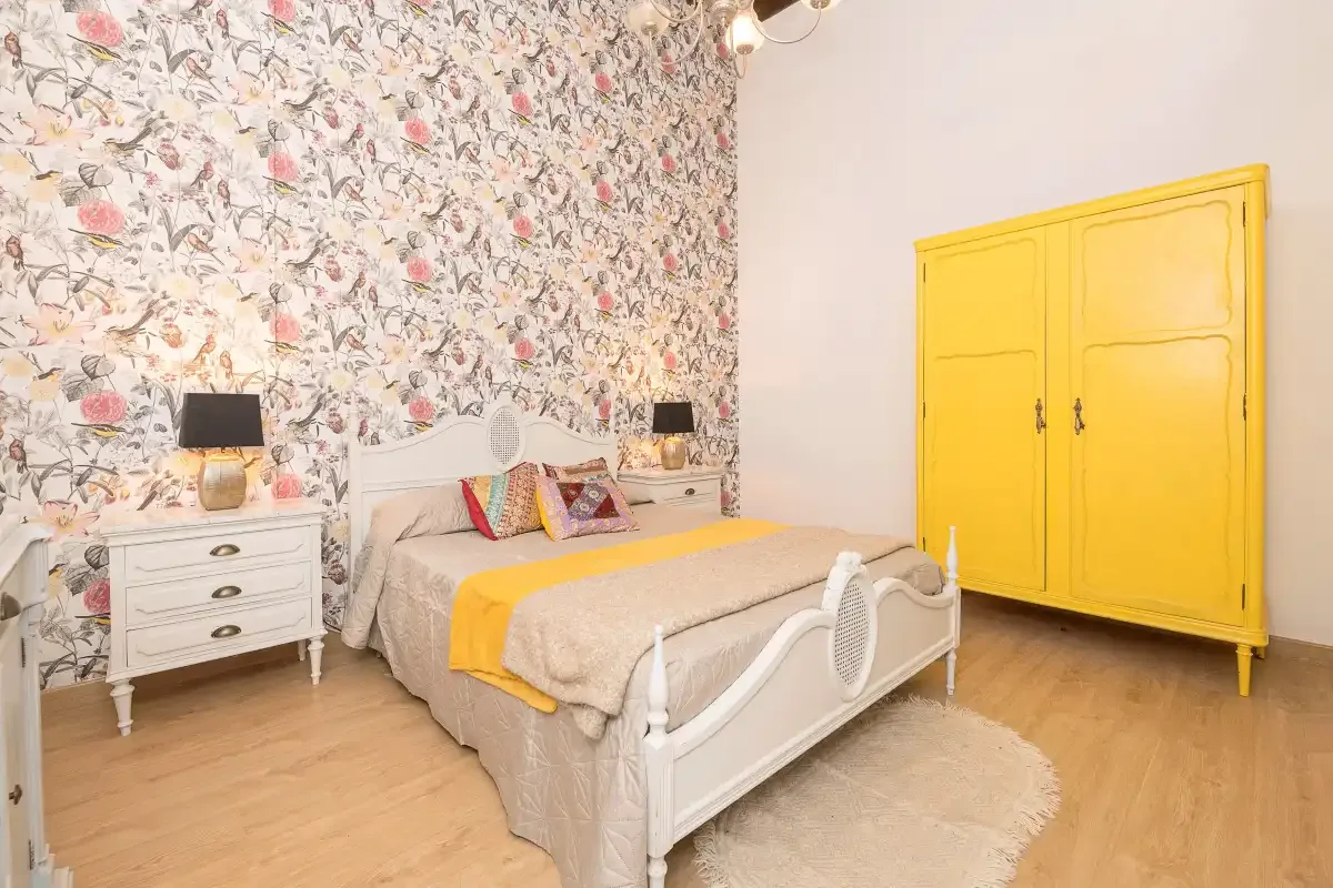 A bedroom with a bed against floral wallpaper and a bright yellow wardrobe.