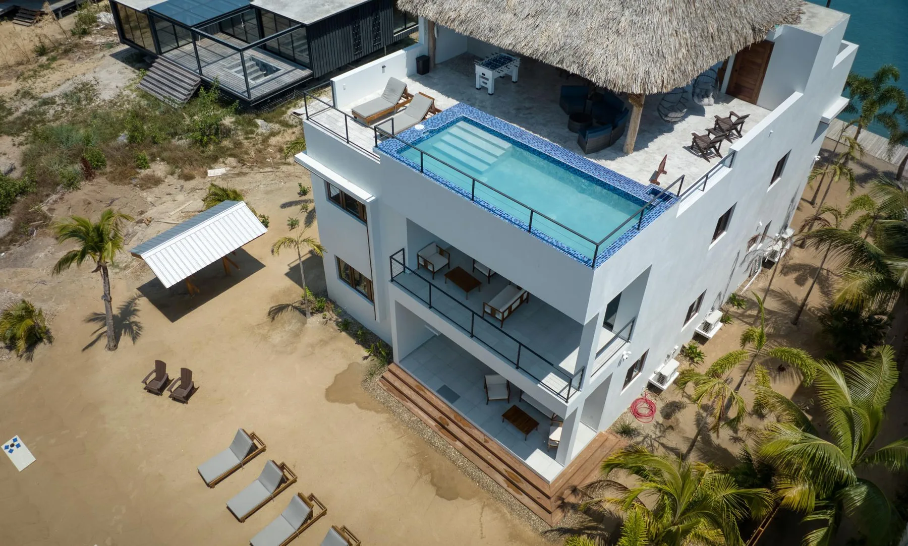 Aerial view of a modern white building with a rooftop pool beside a sandy beach.
