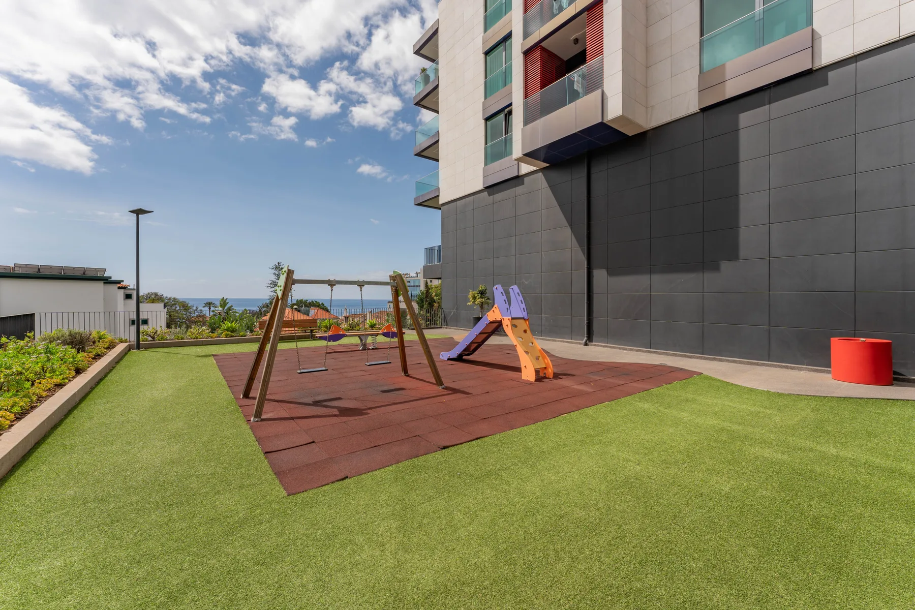 Zona de juegos en una terraza con columpios y un tobogán junto a un edificio moderno.