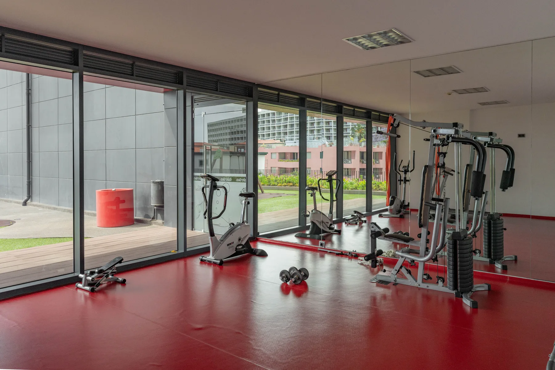 Una sala de ejercicio con equipos y grandes ventanas con vistas a un patio.