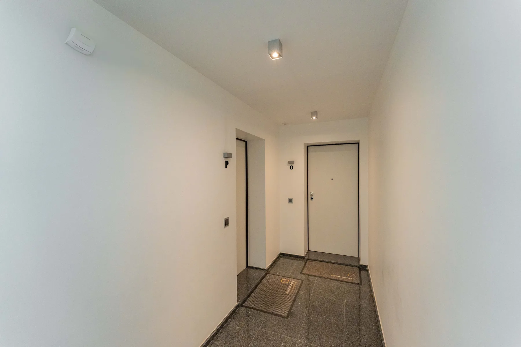Un pasillo interior luminoso con puertas y suelo de baldosas oscuras.