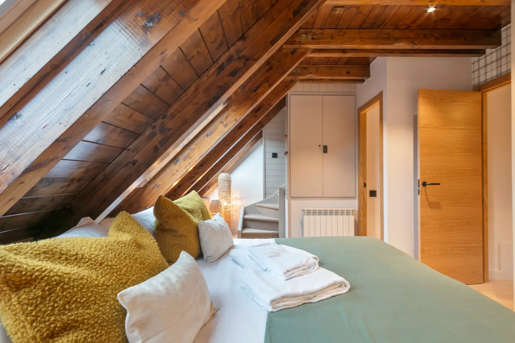 An attic bedroom with a bed beneath a sloped wooden ceiling and a view of nearby buildings.