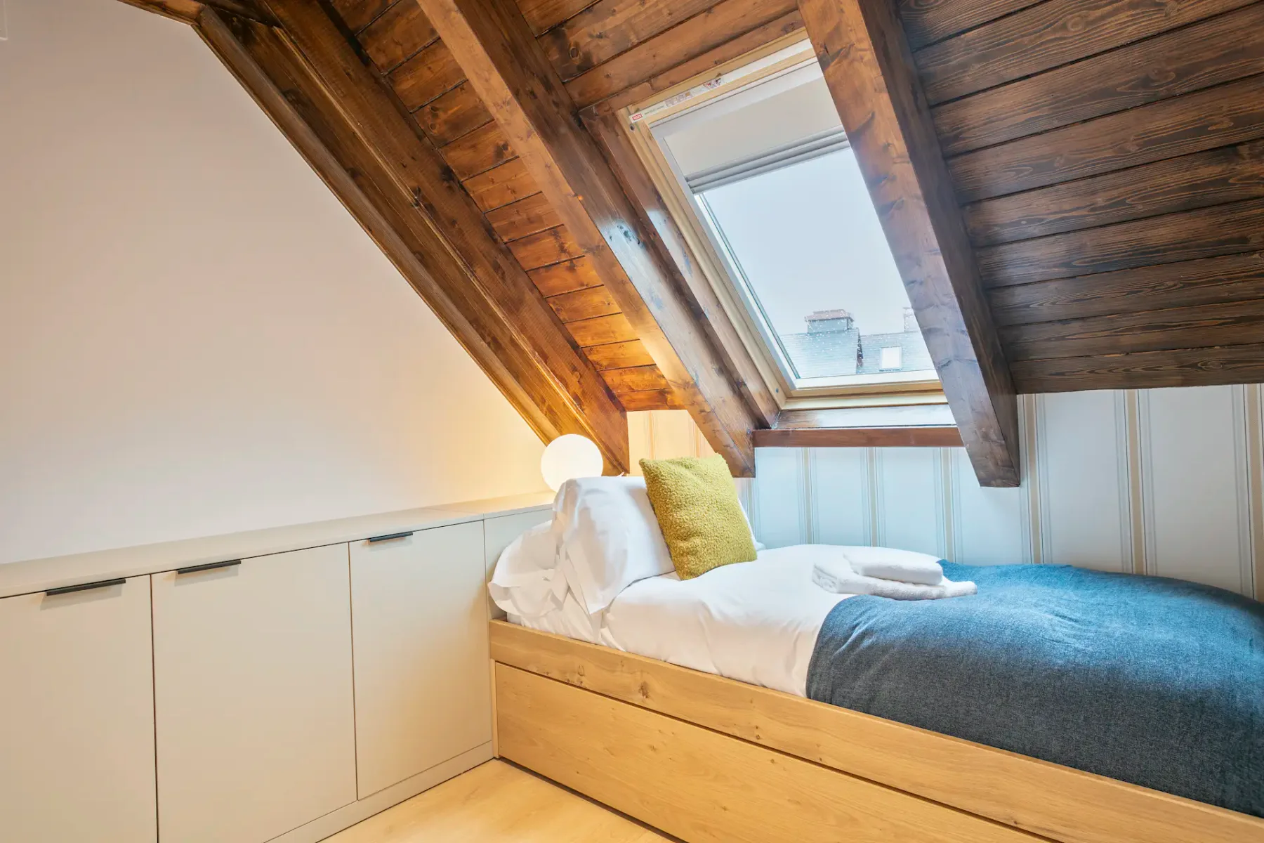 An attic bedroom with a bed beneath a sloped wooden ceiling and skylight.